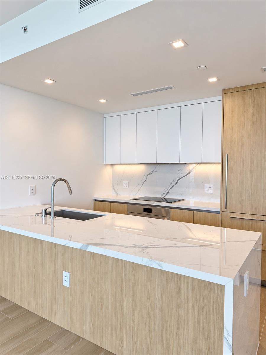 Kitchen featuring a waterfall-edge stone countertop, wood-finish cabinetry, integrated appliances, under-cabinet lighting, and a stone slab backsplash