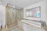 Frameless glass shower enclosure with stone wall tile, built-in shower bench, deep soaking tub with stone surround, and large wall-mounted mirror