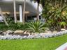 Landscaped garden featuring mature palm trees, decorative rock borders, and manicured turf grass under a curved structural overhang supported by white