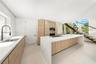 Modern kitchen featuring a waterfall-edge island, light wood-finish cabinetry, stone-style countertops, recessed lighting, and a floating staircase wi