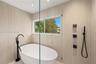 Freestanding white soaking tub with matte black floor-mounted faucet, floor-to-ceiling textured tile wall, frameless glass shower partition, and matte