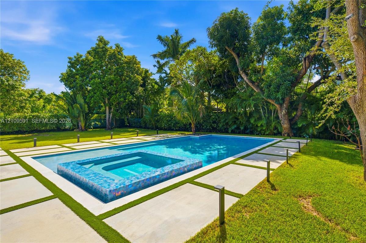 In-ground swimming pool featuring a tiled spillover spa, large-format concrete paver decking, manicured lawn, and mature perimeter landscaping