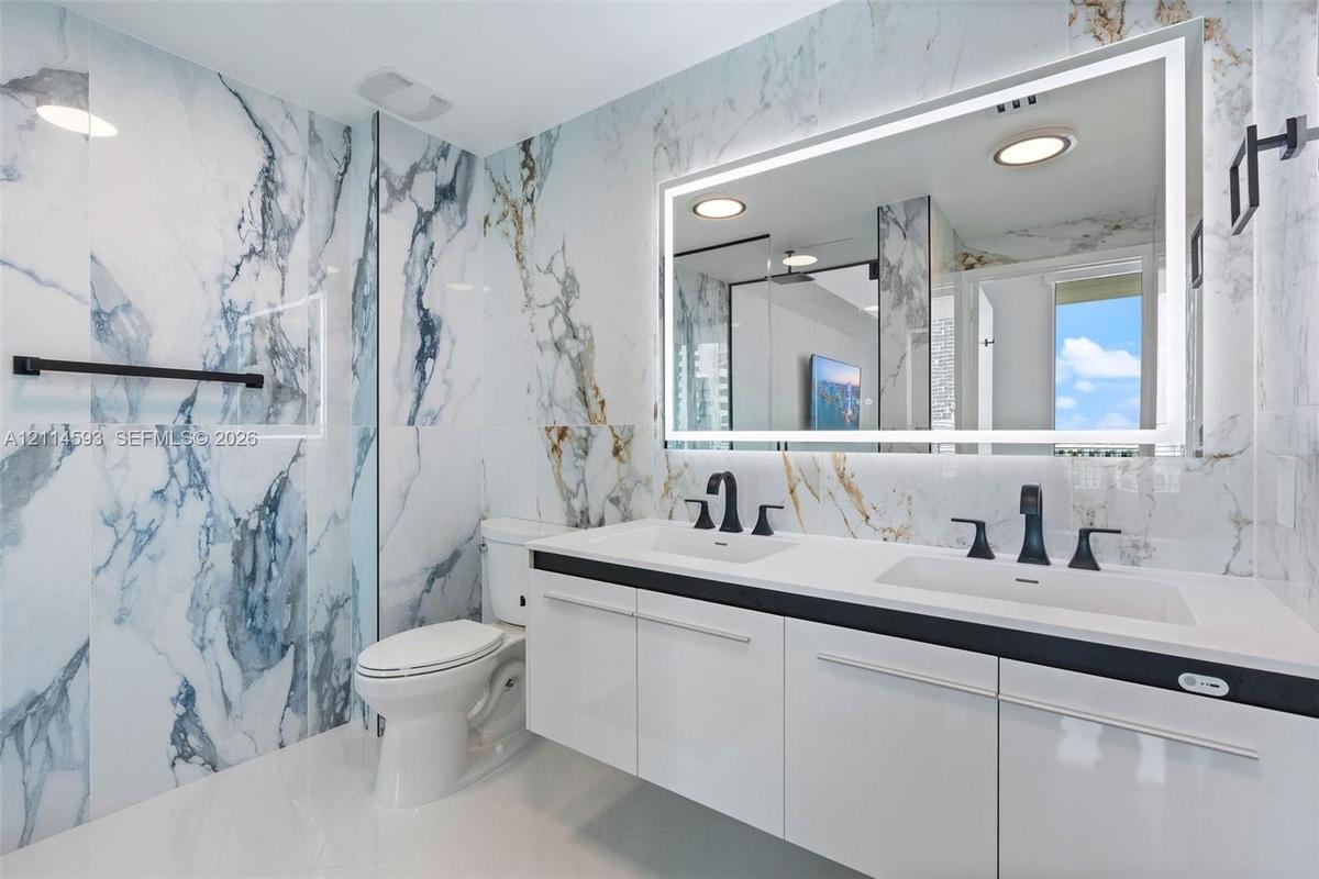 Floating white cabinetry with dual undermount sinks, matte black widespread faucets, large-format stone-finish wall tile, and an integrated LED-backli