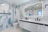Floating white cabinetry with dual undermount sinks, matte black widespread faucets, large-format stone-finish wall tile, and an integrated LED-backli