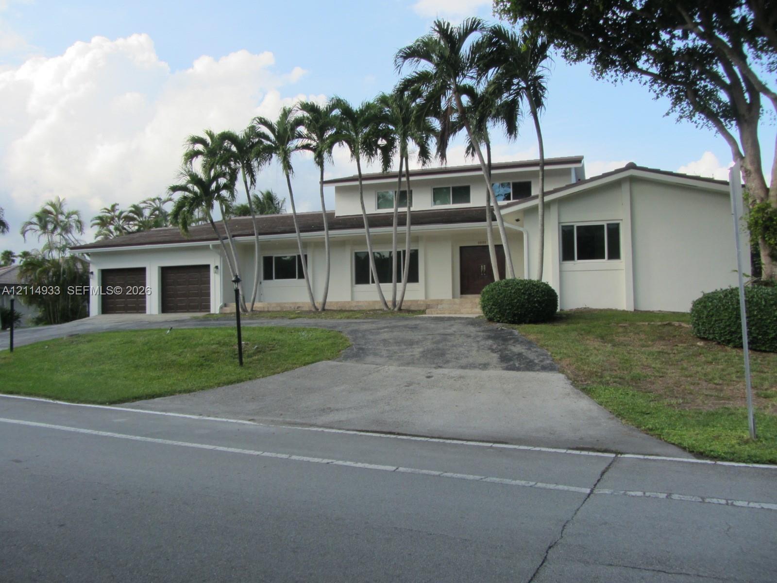 Two-story residence featuring a stucco exterior, multi-car garage with wood-finish doors, expansive driveway, and mature palm tree landscaping