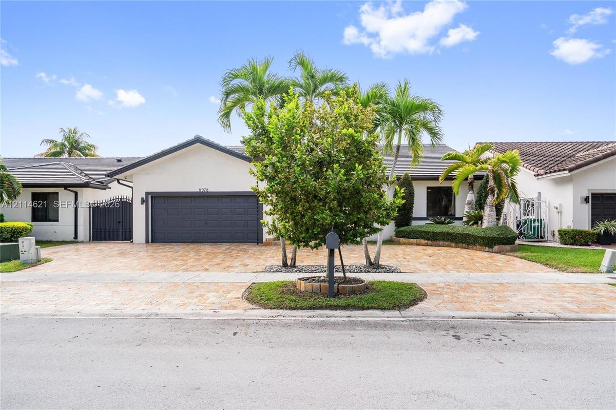 Single-story exterior featuring a light-toned stucco façade, a multi-car garage with a paneled door, a paved driveway, a tile roof, and curated tropic