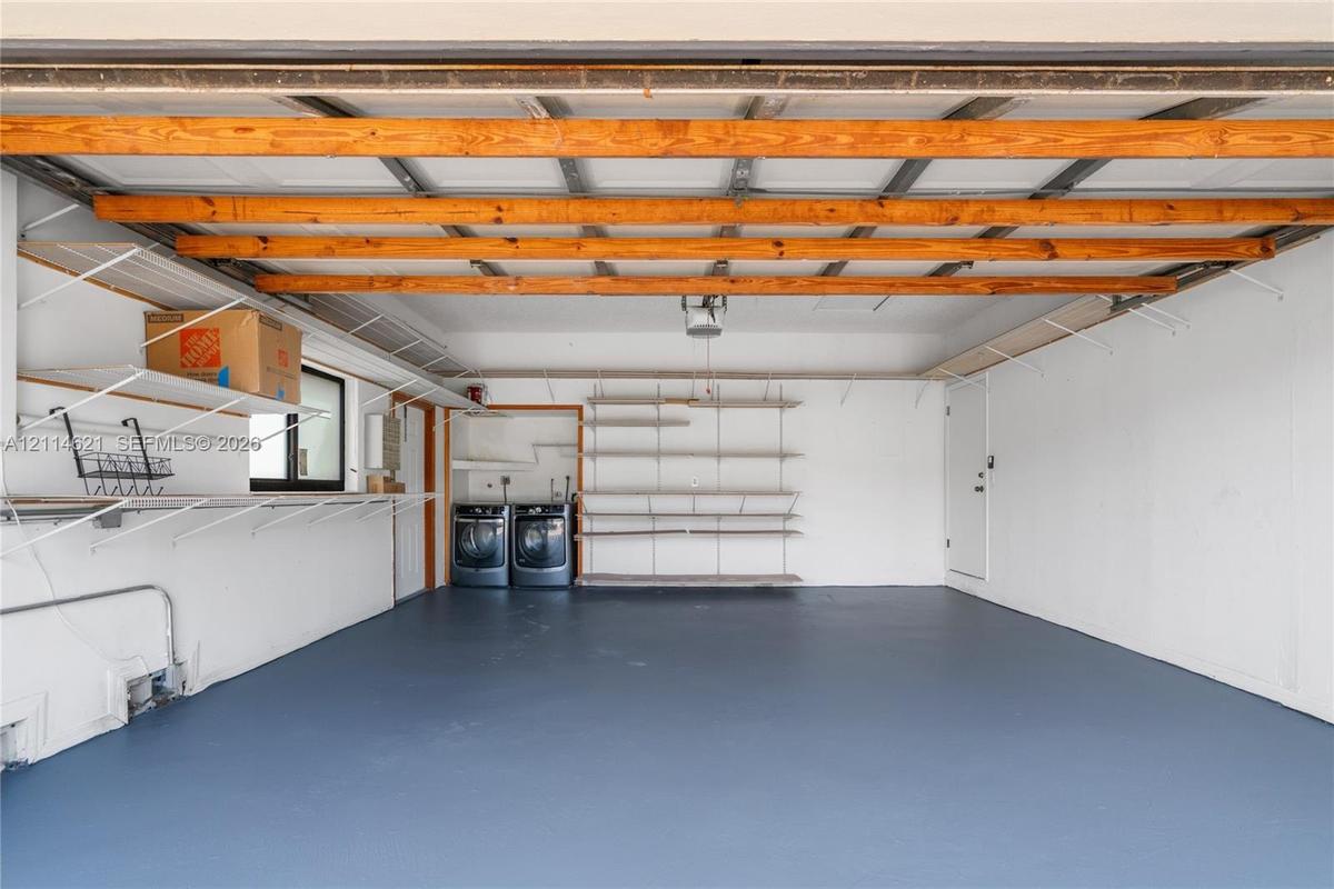 Garage interior featuring exposed wood ceiling beams, painted concrete flooring, mounted metal shelving units, and built-in laundry appliances