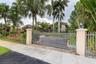 Gated entrance featuring decorative metal fencing, stone-topped stucco pillars with mounted lighting, and a paved driveway surrounded by mature tropic