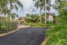 Asphalt driveway with brick-paver accents, stone exterior siding, multi-gabled roofline, metal perimeter fencing, and mature tropical landscaping