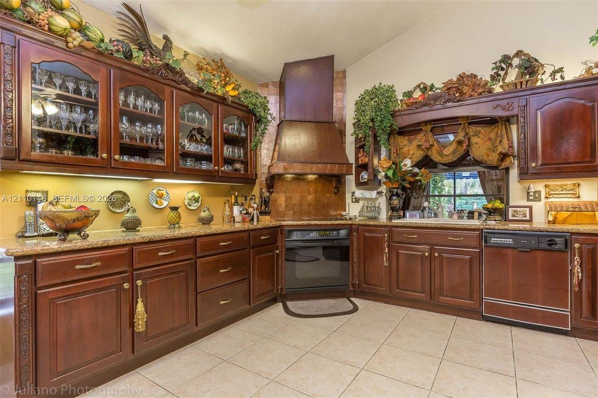 Custom dark wood cabinetry with glass-front display uppers, expansive stone-surface countertops, integrated range hood, built-in oven, and ceramic til
