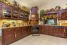Custom dark wood cabinetry with glass-front display uppers, expansive stone-surface countertops, integrated range hood, built-in oven, and ceramic til