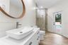 Bathroom featuring a rectangular vessel sink with a chrome faucet, wood-framed circular mirror, walk-in shower with stone-tile surround, wood-finish f