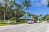 Gated community entrance featuring a teal metal roof structure, mature palm trees, white picket fencing, and manicured perimeter landscaping