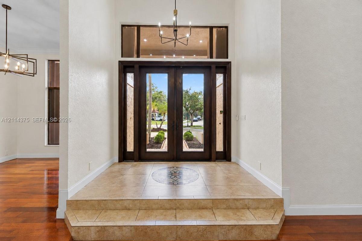 Entryway featuring double glass-paneled doors, decorative tile inlay, stepped tile flooring, transom window, and multi-arm chandelier