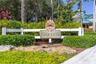 Community signage featuring custom wooden inlay, framed by a white post-and-rail fence, manicured hedges, and mature tropical landscaping