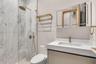 Walk-in shower with vertical stone-finish tiling, brass-toned rainfall shower head, wall-mounted vanity with integrated sink, and illuminated mirrored