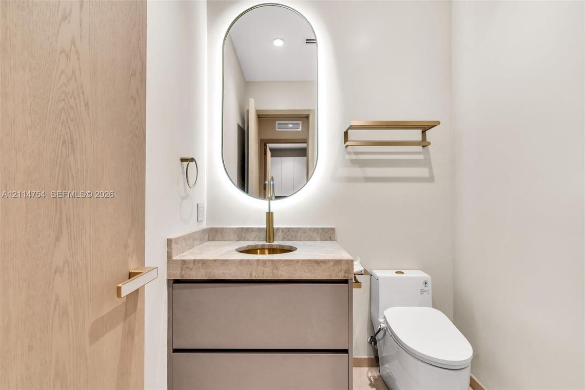 Oval LED-backlit mirror, vanity with integrated stone-finish countertop and brass-tone faucet, soft-close drawers, wall-mounted metallic shelving, and