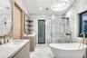 Stone-tiled walls and flooring paired with a freestanding soaking tub, brass fixtures, glass-enclosed walk-in shower, and dual vanities with integrate