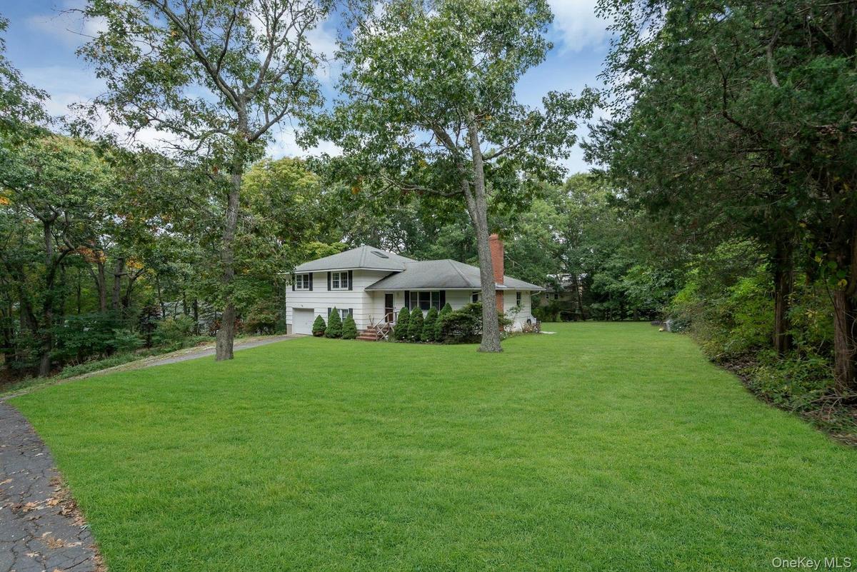 Exterior view of a multi-level residence featuring a light-toned facade, an attached garage, a brick chimney, and an expansive lawn framed by mature t