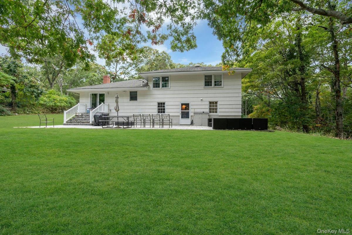 Two-story residence featuring horizontal siding, a stone-paved patio, white railings, and expansive surrounding grounds with mature trees