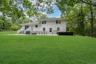 Two-story residence featuring horizontal siding, a stone-paved patio, white railings, and expansive surrounding grounds with mature trees