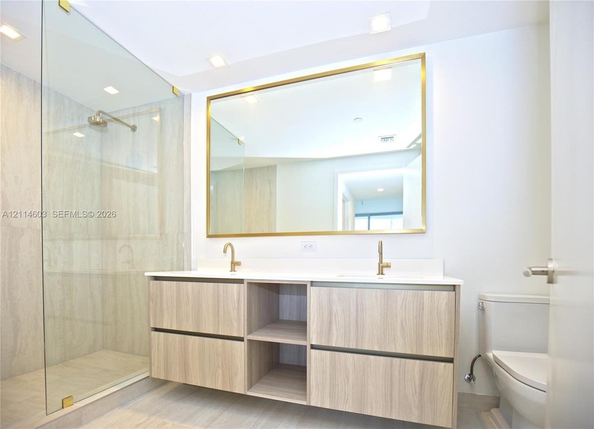 Floating double-vanity with light wood-finish cabinetry, stone-surfaced countertop, glass-enclosed shower with tiled walls, and brass-finish fixtures 