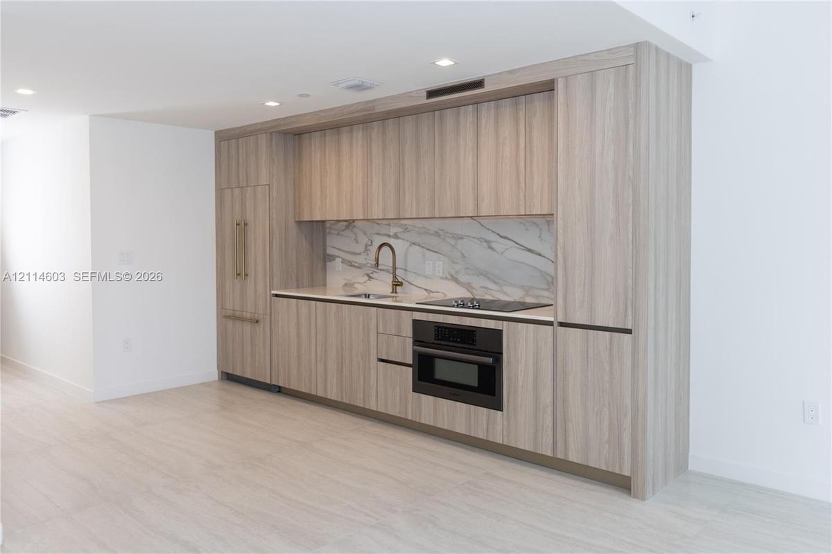 Kitchen featuring flat-panel wood-finish cabinetry, integrated panel-ready refrigerator, stone-patterned backsplash, built-in wall oven, and light-ton