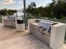 Outdoor kitchen featuring dual stainless steel built-in grills, concrete-surround masonry counters, decorative tile backsplashes, and glass panel rail