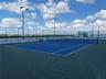 Rooftop tennis court featuring a blue surface with white court markings, perimeter chain-link fencing, and integrated pole-mounted floodlighting