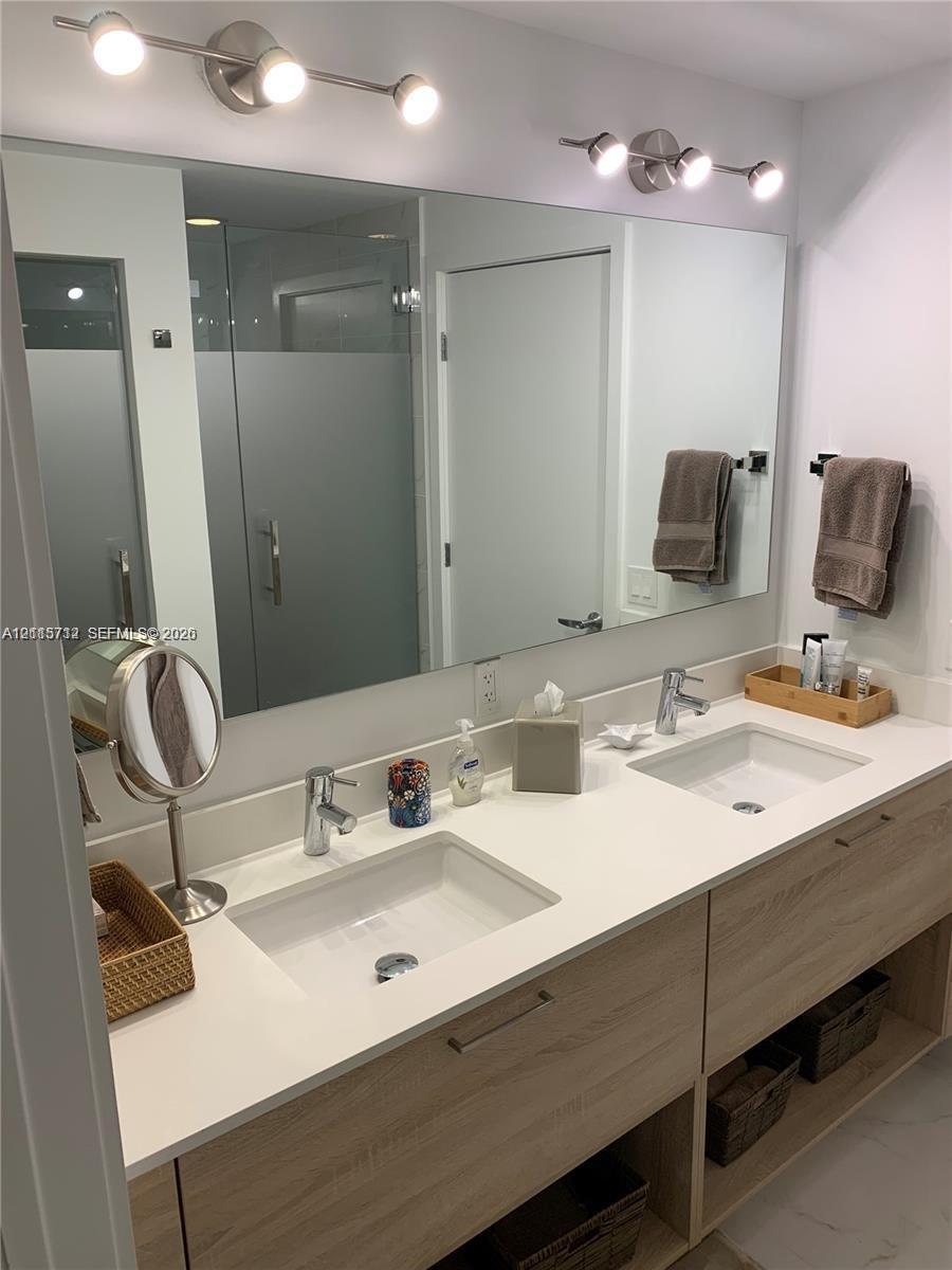 Dual-sink vanity featuring a light wood-finish cabinet with open shelving, a white surface, undermount rectangular sinks, and brushed metal hardware