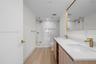 Dual-sink vanity with quartz countertop, light wood-finish cabinetry, brass-toned fixtures, rectangular mirror with metallic frame, and a glass-enclos