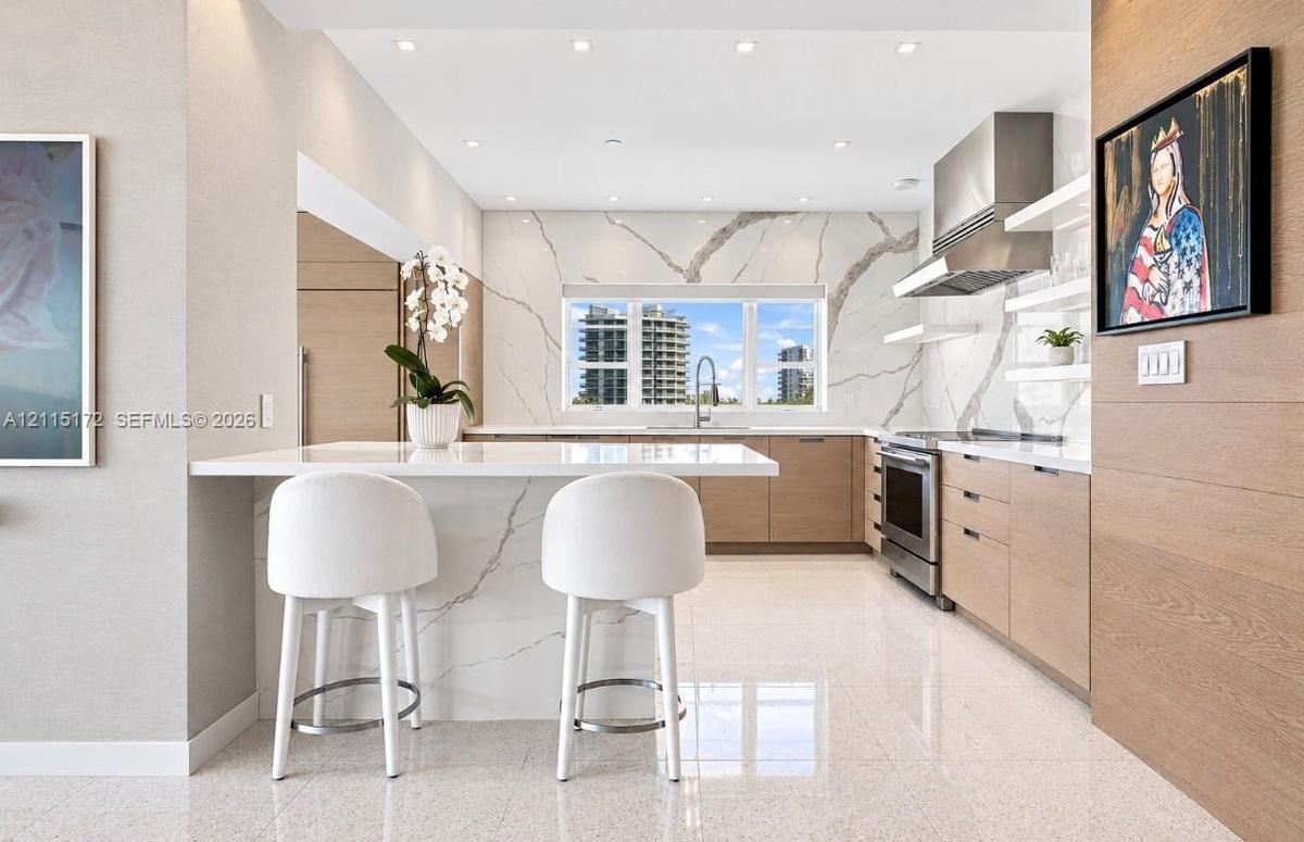 Kitchen featuring a waterfall-edge stone island, veined stone backsplash, stainless steel range hood, flat-panel wood-finish cabinetry, and recessed c