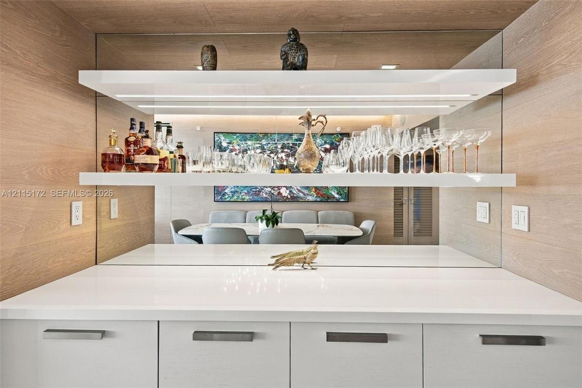 Custom built-in bar featuring white cabinetry, polished solid-surface countertop, mirrored backsplash, and tiered floating shelving with integrated LE