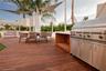 Outdoor terrace featuring wood-finish decking, a built-in stainless steel grill station, white structural shade sails, and integrated landscaping