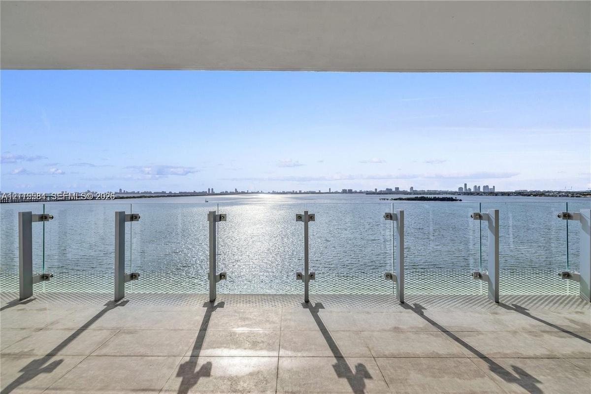 Covered terrace featuring large-format stone flooring and a glass-panel railing overlooking the water and distant cityscape