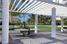 Outdoor space featuring a white slatted pergola, cylindrical support columns, stone paver flooring, a permanent stone table, and a tiered lawn area