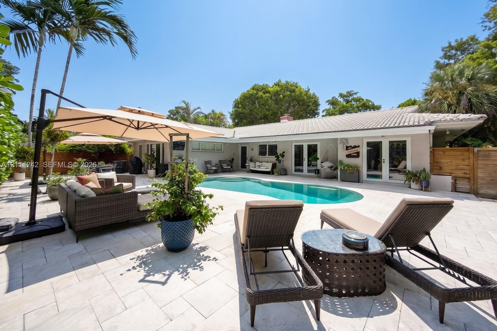 Backyard featuring a custom-shaped swimming pool, extensive light-toned stone hardscaping, multiple glass French door entries, and mature tropical pal
