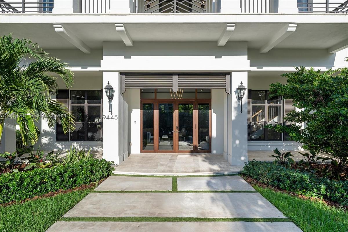 Covered entryway featuring a double-door glass entrance, concrete paver walkway with turf accents, dual exterior wall sconces, and architectural suppo