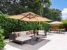 Outdoor patio area featuring light-toned stone pavers, tall perimeter privacy hedging, mature tropical foliage, and natural wood fencing