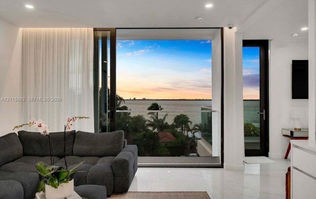 Floor-to-ceiling sliding glass doors, glass balcony railing, recessed lighting, and white stone-finish flooring with water horizon backdrop