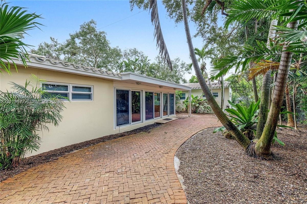 Stucco exterior featuring a curved brick walkway, series of glass sliding doors, and a tile roof set among mature tropical landscaping