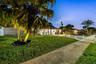 Exterior facade featuring a concrete driveway, manicured lawn, mature palm trees, white vinyl privacy fencing, and exterior landscape lighting