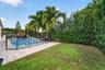 In-ground swimming pool with tile surround, black perimeter safety fencing, mature palm trees, and dense natural foliage hedging