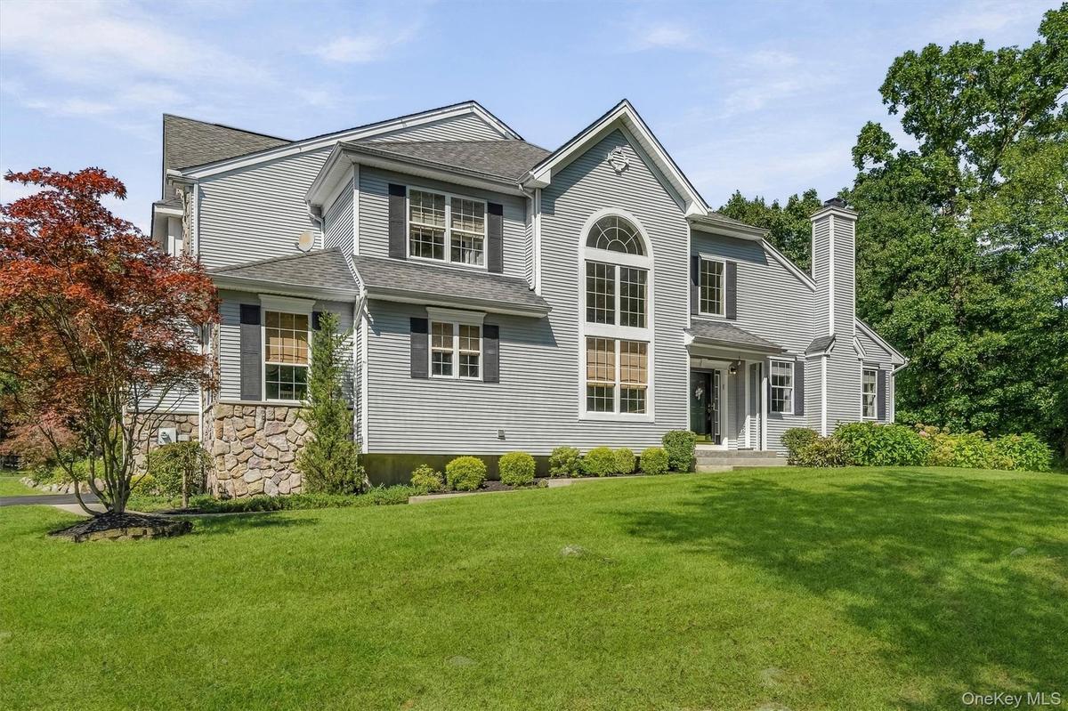 Expansive front lawn with mature landscaping, a stone veneer  foundation, multiple windows with black shutters, an arched transom window, and a promin