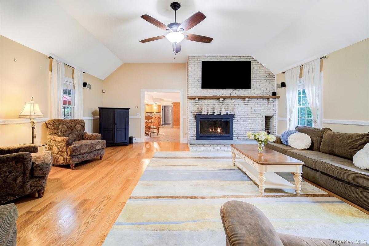 Vaulted ceiling, ceiling fan with integrated lighting, natural wood-finish flooring, white-painted brick fireplace with wood mantel, and multiple wind
