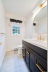 First floor 1/2 bath features wood-finish vanity with gold-tone hardware, white stone countertop, round gold-tone framed mirror, tiled flooring and wh