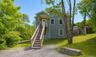 Front entry with covered stoop, exterior staircase, gravel driveway and mature shade trees.