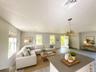 Virtually staged. Living room with three windows and chandelier, viewed from the kitchen island.