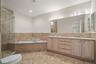 Travertine tile flooring, built-in bathtub, glass-enclosed shower, dual vanity with undermount sinks, and a large wall-mounted mirror