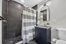 Shower with dark tile surround and pebble floor, dark vanity cabinet with light countertop, arched mirror, and contemporary light fixture